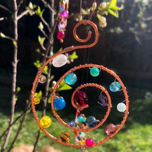 Könnte beinhalten: Ein spiralförmiger Sonnenfänger aus Kupferdraht, verziert mit bunten Glasperlen. Das Spiraldesign weist eine Vielzahl von Schmuckperlen in Blau-, Grün-, Rot- und Gelbtönen auf. Ein kleinerer Perlenstrang hängt darunter. Der Sonnenfänger ist an einem dünnen Draht aufgehängt.