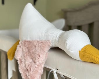 Könnte beinhalten: Ein weißes Kissen in Form einer Gans mit einem flauschigen rosa Flügel und einem gelben Schnabel, das auf einem weißen Kinderbett-Stoßfänger liegt.