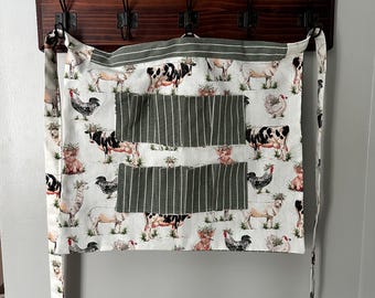 May include: A white apron with a farm animal print, including cows, pigs, chickens, and sheep. The apron has two large green striped pockets and brown straps.