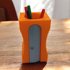 May include: A bright orange pencil sharpener shaped desk organizer with a light blue front panel featuring a small plus sign symbol. The top opening contains three thin pens in red, green, and black colors, standing upright inside the container.
