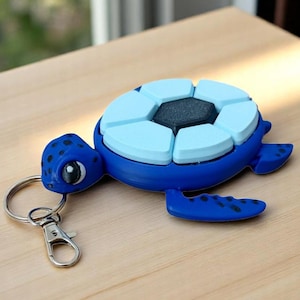 May include: A blue sea turtle keyring featuring a dark blue head with black spots and a white eye. The shell is composed of six light blue segments surrounding a central dark grey hexagonal piece. A silver metal split ring and lobster clasp are attached.