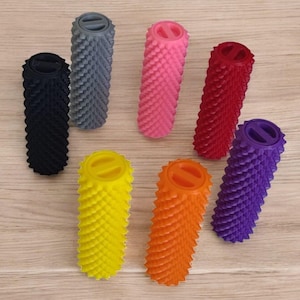 Op de afbeelding: Een verzameling van zeven cilindrische massagerollers in diverse kleuren, waaronder zwart, grijs, roze, rood, geel, oranje en paars. Elke roller heeft een getextureerd oppervlak met stekels en een platte bovenkant met een horizontale gleuf.