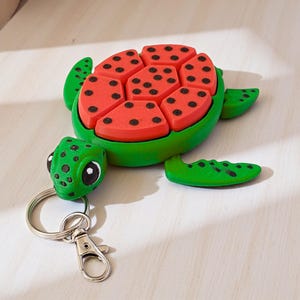 May include: A turtle-shaped keychain featuring a green head and limbs with black spots, and a shell designed to resemble a watermelon slice with a bright red center, black seeds, and a green border, attached to a silver metal split ring and clasp.