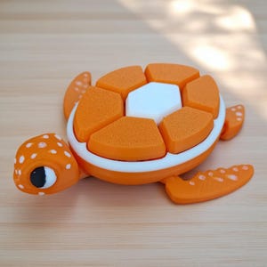 May include: A small, articulated sea turtle figurine featuring a bright orange body with white polka dots on the head and flippers. The shell is composed of segmented orange pieces surrounding a central white hexagonal plate, all bordered by a white rim.