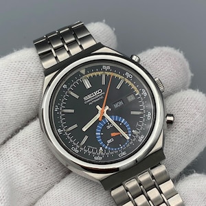 May include: A Seiko Chronograph Automatic watch with a black dial, silver-tone stainless steel link bracelet, and a day-date window. The watch face features a bright orange second hand, a blue and white sub-dial, and white hour markers.