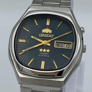以下が含まれることがあります： ダークブルーのテクスチャ文字盤を備えたシルバーカラーのステンレススチール製腕時計。ゴールドカラーのインデックスと針、3時位置の曜日・日付表示窓、3つの星と「21 JEWELS」の文字が記されたオリエントのロゴが特徴です。