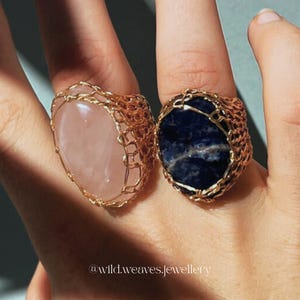 May include: Two handmade rings with large, oval gemstones. One ring features a light pink stone, the other a dark blue stone. Both stones are encased in a woven gold-colored wire setting. The rings are displayed on a finger.