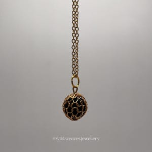 May include: A gold-tone necklace with a chain and a spherical pendant. The pendant has a black sphere within a gold wire cage. The necklace is set against a plain backdrop. The text "@wild.weaves.jewellery" is visible at the bottom of the image.