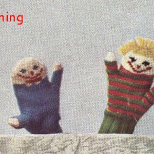 May include: Two children wearing knit hats, one blue and one green, are positioned between four knitted finger puppets. The puppets feature smiling faces with red mouths and yellow hair. The text "heresthething" is printed in red above the blue hat.
