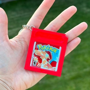 Puede incluir: Un llavero rectangular de color rojo brillante modelado como un cartucho de videojuego portátil clásico. La etiqueta central presenta el logotipo de Pokémon, una ilustración del personaje Charizard y un símbolo de clasificación ESRB E.