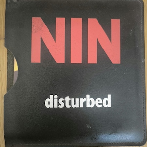 May include: Black vinyl CD case with the red letters "NIN" and the word "disturbed" in white. The case has a pocket on the left side and is likely used to store CDs.