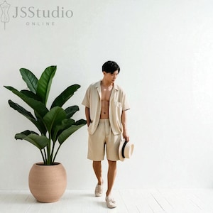May include: A beige linen short-sleeve shirt and shorts set. The model is holding a straw hat and wearing beige shoes. A large potted plant is to the left of the model. The text "JSStudio ONLINE" is in the upper left corner.