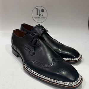 May include: A pair of black leather dress shoes with decorative perforations and a white and brown stitched border. The shoes have laces and a low heel. The Leather Lords logo is visible in the background.