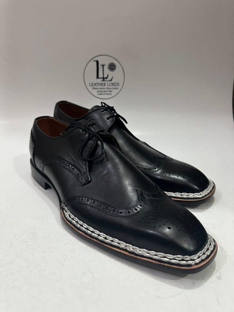May include: A pair of black leather dress shoes with decorative perforations and a white and brown stitched border. The shoes have laces and a low heel. The Leather Lords logo is visible in the background.