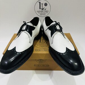 Può includere: Un paio di scarpe eleganti in pelle bicolore, nere e bianche. Le scarpe presentano un design a punta di ala con perforazioni decorative e lacci neri. Sono esposte su una scatola marrone con la scritta "LEATHER LORDS".