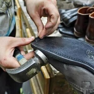 May include: A cobbler uses a hammer to repair a black leather shoe sole. The close-up shot shows the craftsman's hands working on the shoe, with other shoes visible in the background. The hammer is silver and the sole is black.