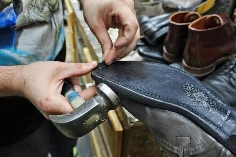 May include: A cobbler uses a hammer to repair a black leather shoe sole. The close-up shot shows the craftsman's hands working on the shoe, with other shoes visible in the background. The hammer is silver and the sole is black.