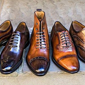 May include: A collection of men's leather dress shoes in various shades of brown and black. Styles include oxfords, wingtips, and a high-top boot, all with laces. The shoes are arranged on a stone surface, showcasing their polished finish.