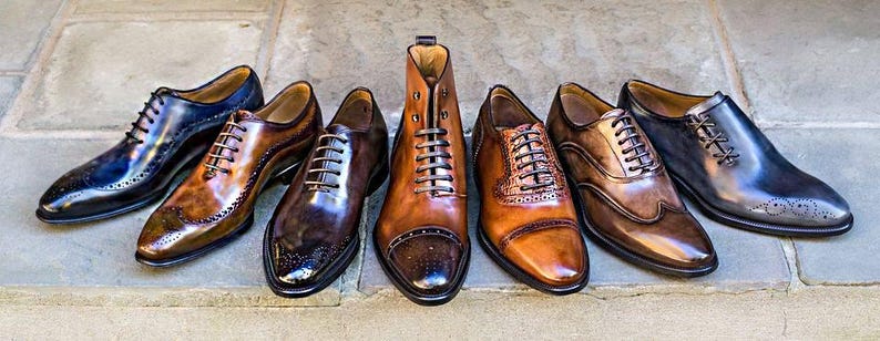 May include: A collection of men's leather dress shoes in various shades of brown and black. Styles include oxfords, wingtips, and a high-top boot, all with laces. The shoes are arranged on a stone surface, showcasing their polished finish.