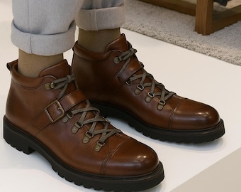 May include: Brown leather boots with dark green laces and a buckle closure. The boots have a lug sole and are displayed on a white surface. The boots are from Leather Lords, handcrafted.