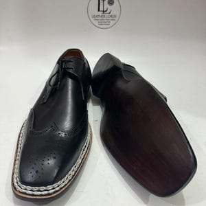 May include: Black leather dress shoes with decorative perforations and a white stitched border. The shoes have a classic lace-up design and a dark brown sole. The image shows the shoes from a low angle, highlighting their details.