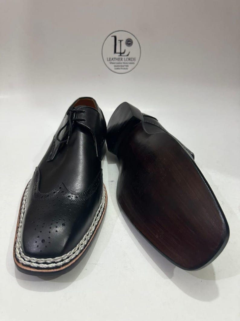 May include: Black leather dress shoes with decorative perforations and a white stitched border. The shoes have a classic lace-up design and a dark brown sole. The image shows the shoes from a low angle, highlighting their details.