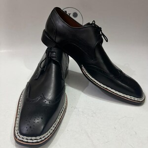 May include: A pair of black leather dress shoes with a wingtip design. The shoes have a stitched white and brown border around the soles and are laced up. The shoes are displayed on a white surface.