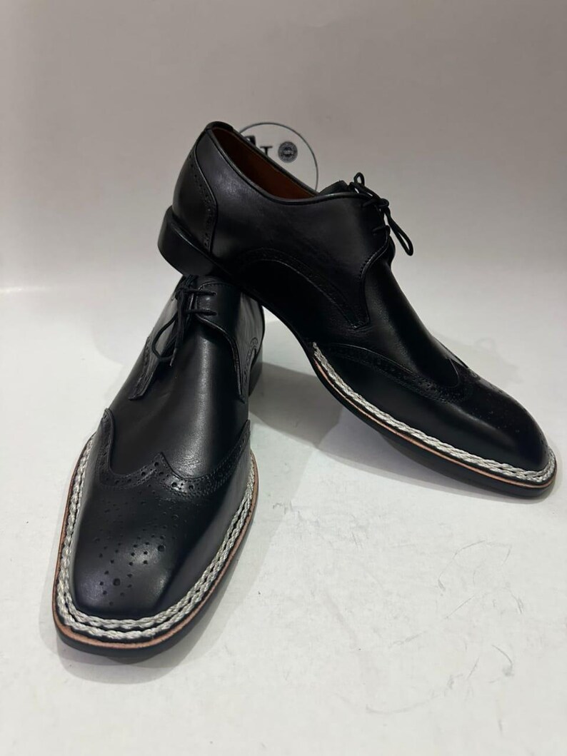 May include: A pair of black leather dress shoes with a wingtip design. The shoes have a stitched white and brown border around the soles and are laced up. The shoes are displayed on a white surface.