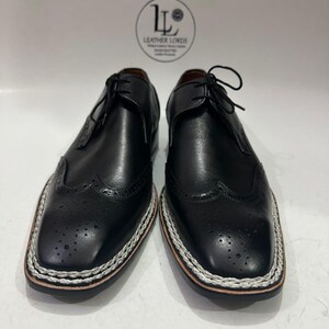 May include: A pair of black leather dress shoes with a square toe and decorative perforations. The shoes have a white and silver stitched border around the sole and are laced up. The brand logo "Leather Lords" is visible in the background.