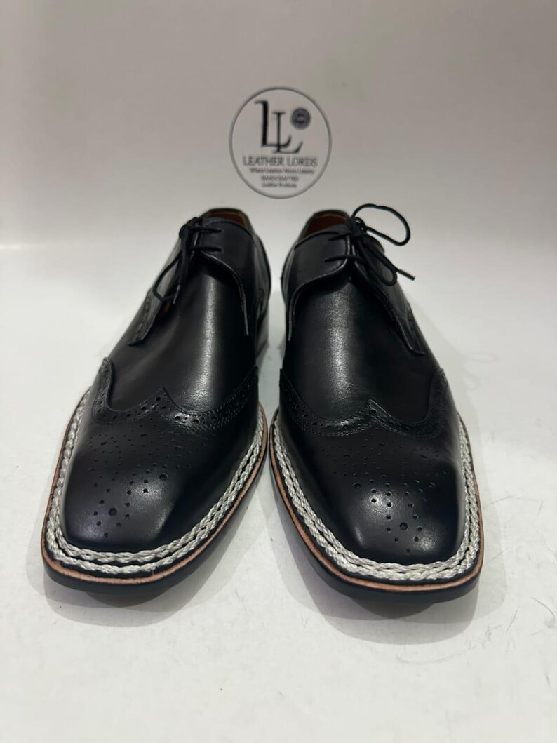 May include: A pair of black leather dress shoes with a square toe and decorative perforations. The shoes have a white and silver stitched border around the sole and are laced up. The brand logo "Leather Lords" is visible in the background.