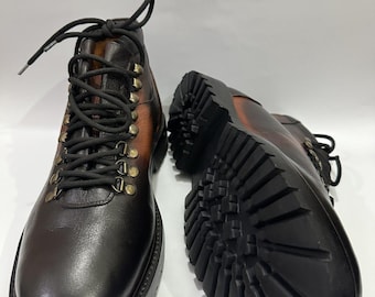 May include: A pair of brown leather boots with black laces and brass eyelets. The boots have a rugged, lug sole and a two-tone color scheme, transitioning from dark brown to a lighter shade at the top.