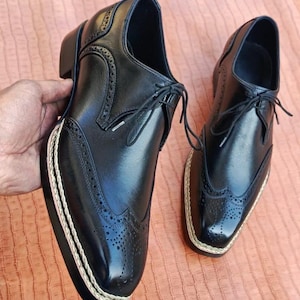 May include: Pair of black leather dress shoes with brogue detailing and a cream-colored rope trim around the sole. The shoes have a classic lace-up design and a small heel. The shoes are displayed on a peach-colored surface.