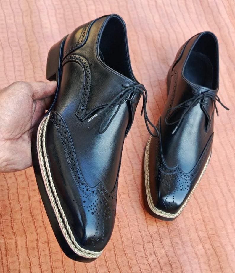 May include: Pair of black leather dress shoes with brogue detailing and a cream-colored rope trim around the sole. The shoes have a classic lace-up design and a small heel. The shoes are displayed on a peach-colored surface.