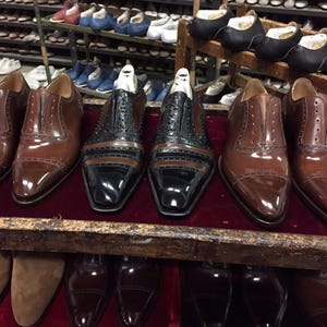 May include: A collection of leather dress shoes in various stages of construction. The shoes are primarily brown and black, with intricate stitching and detailing. Some shoes have wooden shoe forms inside. The background shows shelves of other shoes.