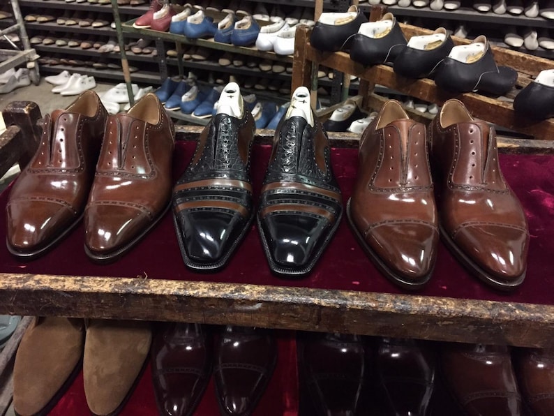 May include: A collection of leather dress shoes in various stages of construction. The shoes are primarily brown and black, with intricate stitching and detailing. Some shoes have wooden shoe forms inside. The background shows shelves of other shoes.