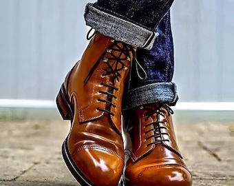 May include: A pair of polished tan leather lace-up boots with a cap toe design, featuring dark brown laces and metal eyelets. The boots have a stacked heel and are worn with dark blue cuffed denim jeans that show a grey interior lining.