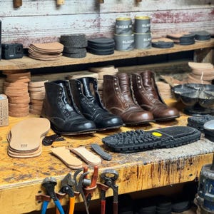 May include: A workbench displays leather boots in various stages of construction. Black and brown leather boots are lined up, alongside tools, leather soles, and materials. Shelves above hold more supplies, including cans of black shoe polish.