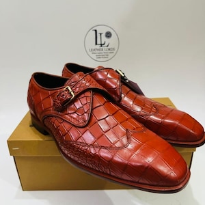 May include: A pair of reddish-brown leather dress shoes with a crocodile skin pattern. The shoes have a buckle closure and are displayed on a cardboard box. The brand logo "Leather Lords" is visible in the background.