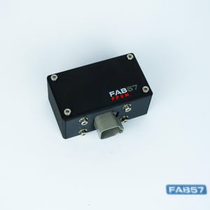 Puede incluir: Un módulo de control electrónico rectangular de color negro con cuatro tornillos plateados en la superficie superior y un puerto de conexión de plástico gris en el lateral. El texto FAB57 FPCM está impreso en blanco y rojo en la carcasa.