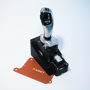 May include: A car gear shift lever with a black and silver design. The lever has a black top with white lettering indicating gear positions. A black base and a rust-colored card with "FAB57" are also visible.