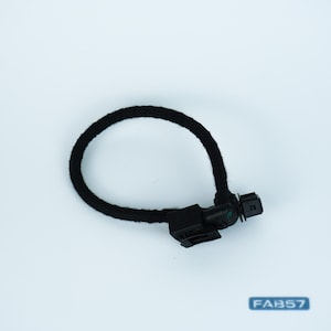 May include: A black automotive electrical sensor component featuring a circular, fabric-wrapped wire harness loop connected to a rigid plastic connector housing. The assembly includes a small black plug with a square port and a secondary smaller connector.
