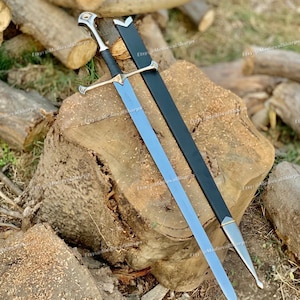 Peut inclure: Une épée en acier à double tranchant avec une poignée gainée de cuir noir, un pommeau argenté et une garde transversale. L'épée est accompagnée d'un fourreau noir doté de garnitures métalliques argentées à la chape et à la bouterolle.