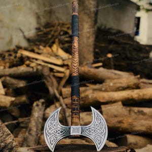 May include: A double-bitted battle axe featuring a silver-colored metal head with intricate Celtic knotwork engravings. The long wooden handle is carved with geometric patterns and wrapped with black cord at the top and middle sections.