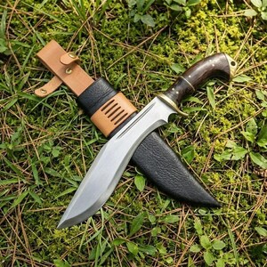 Puede incluir: Un cuchillo de hoja grande y curvada con mango de madera oscura y detalles en latón, en una funda de cuero negro. El cuchillo tiene una trabilla de cinturón de cuero marrón claro, sobre hierba y musgo verdes.
