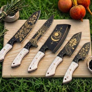 Könnte beinhalten: Ein Set aus fünf Küchenmessern mit schwarzen Klingen, die goldfarbene Gravuren wie einen Phönix, einen Baum, Sonne und Mond, ein Eichenblatt und florale Muster aufweisen. Jedes Messer hat einen hellen, strukturierten Griff und eine Metallzwinge.