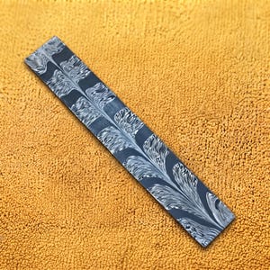 May include: A rectangular metal bar featuring a dark gray surface with a repeating white feather-like pattern. The material displays a distinct layered appearance with flowing, organic lines that create a symmetrical, branching visual effect.
