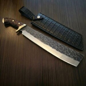 May include: A large, hand-forged knife with a textured blade and a wooden handle. The knife is accompanied by a black leather sheath with a crocodile pattern. The blade is approximately 12 inches long.