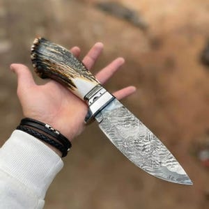 Könnte beinhalten: Ein feststehendes Messer mit einem strukturierten, braun-cremefarbenen Geweihgriff, einem polierten Metallbeschlag und einer scharfen, silberfarbenen Klinge, die ein ausgeprägtes, wellenförmiges Damaststahlmuster auf der gesamten Metalloberfläche aufweist.
