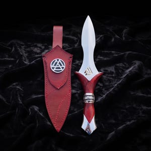 May include: A double-edged dagger with a polished silver blade, a red wood handle, and a silver pommel. The blade features a gold-colored Valknut symbol. It is accompanied by a red leather sheath adorned with a circular silver Valknut emblem.