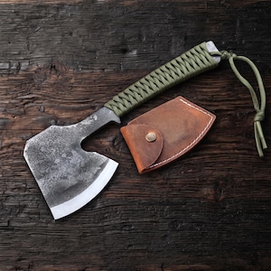 Peut inclure: Une hachette en acier forgé à la main avec une finition sombre et texturée et un tranchant affûté. Le manche est enveloppé de paracorde vert olive avec une dragonne assortie. Un étui en cuir marron avec des coutures blanches est posé à côté.
